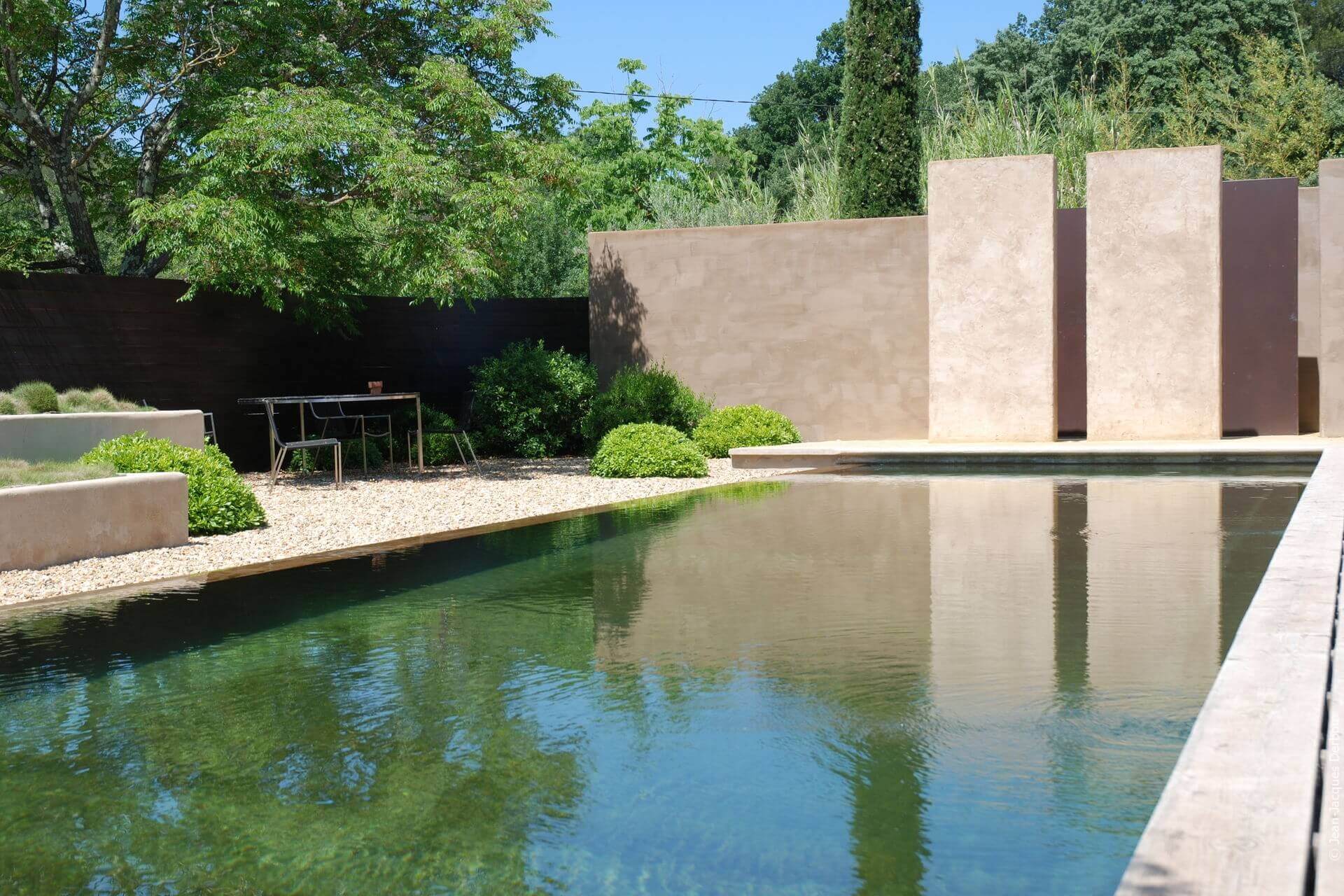 Mobilier contemporain à l’ombre, au bord de la piscine biologique, gravier, mur enduit pigment naturel.