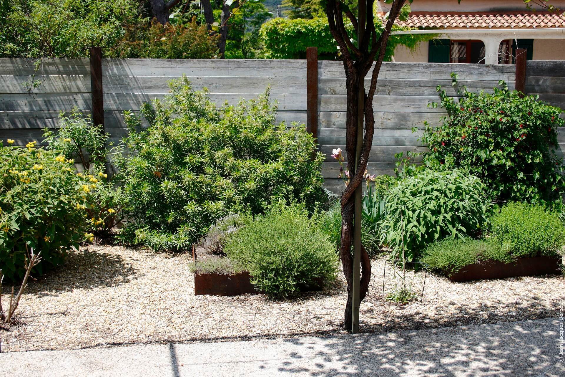 Clôture de jardin en bois recyclé et poteaux en métal brut, jardin en carré, jardinière en métal brut, gravier, plantes de jardin sans arrosage.