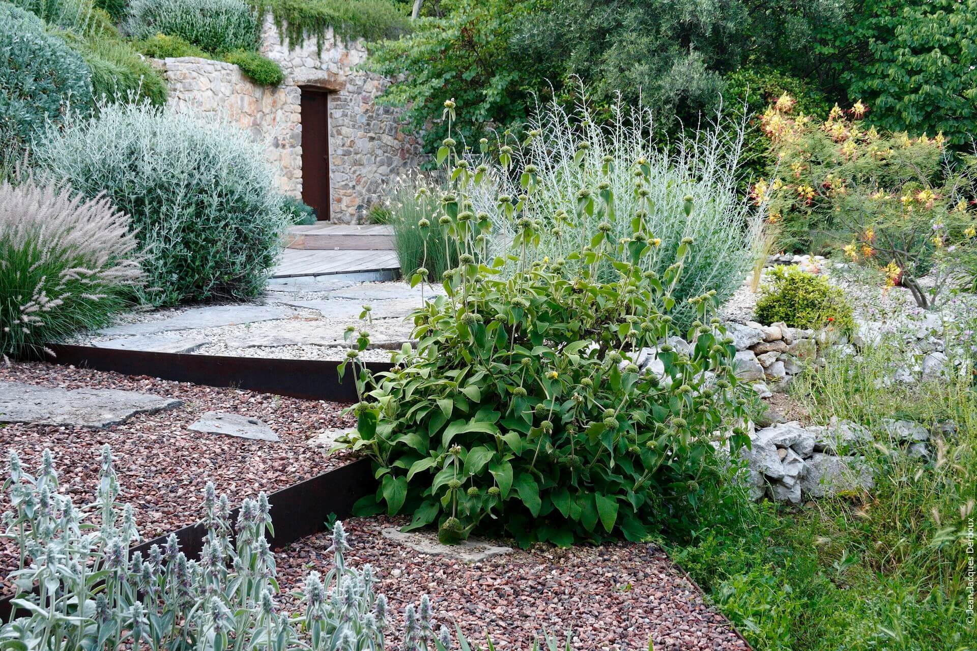 Jardin sans arrosage, ambiance naturelle, muret en pierre, terrasse en bois, grande dalle naturelle, gravier, contre marche en métal brut, plantes de méditerranée.